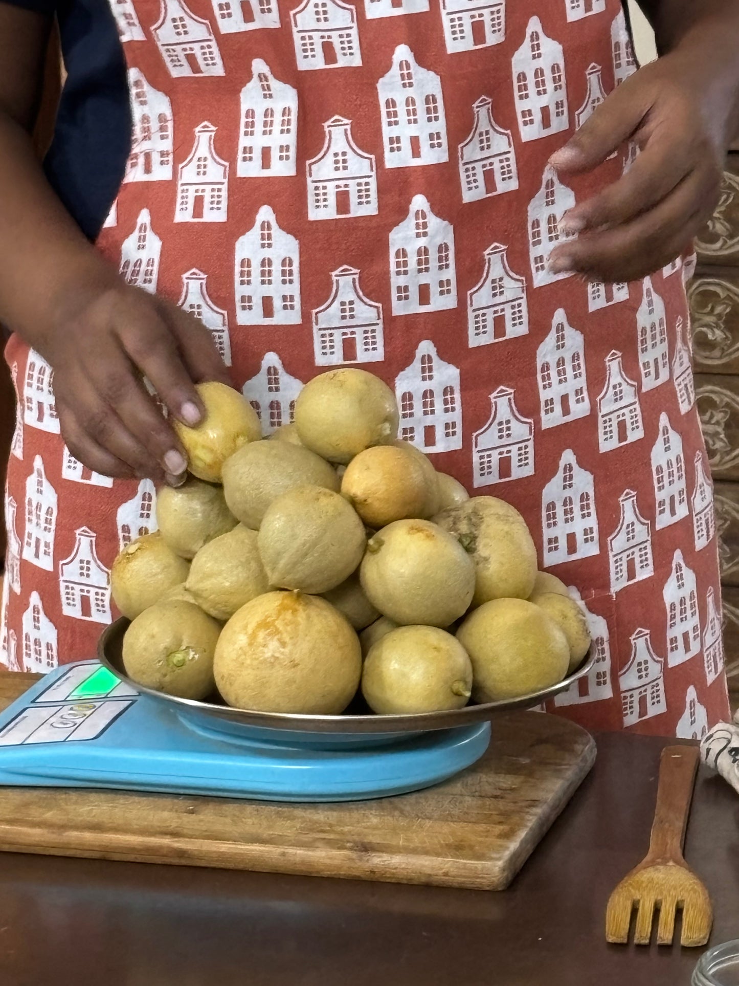 A pile of round, pale yellow fruits, likely papayas or guavas, sits on a metal tray on a blue digital scale. A person wearing a red apron with a white house pattern is visible from the chest down, with their hands reaching toward the fruit.