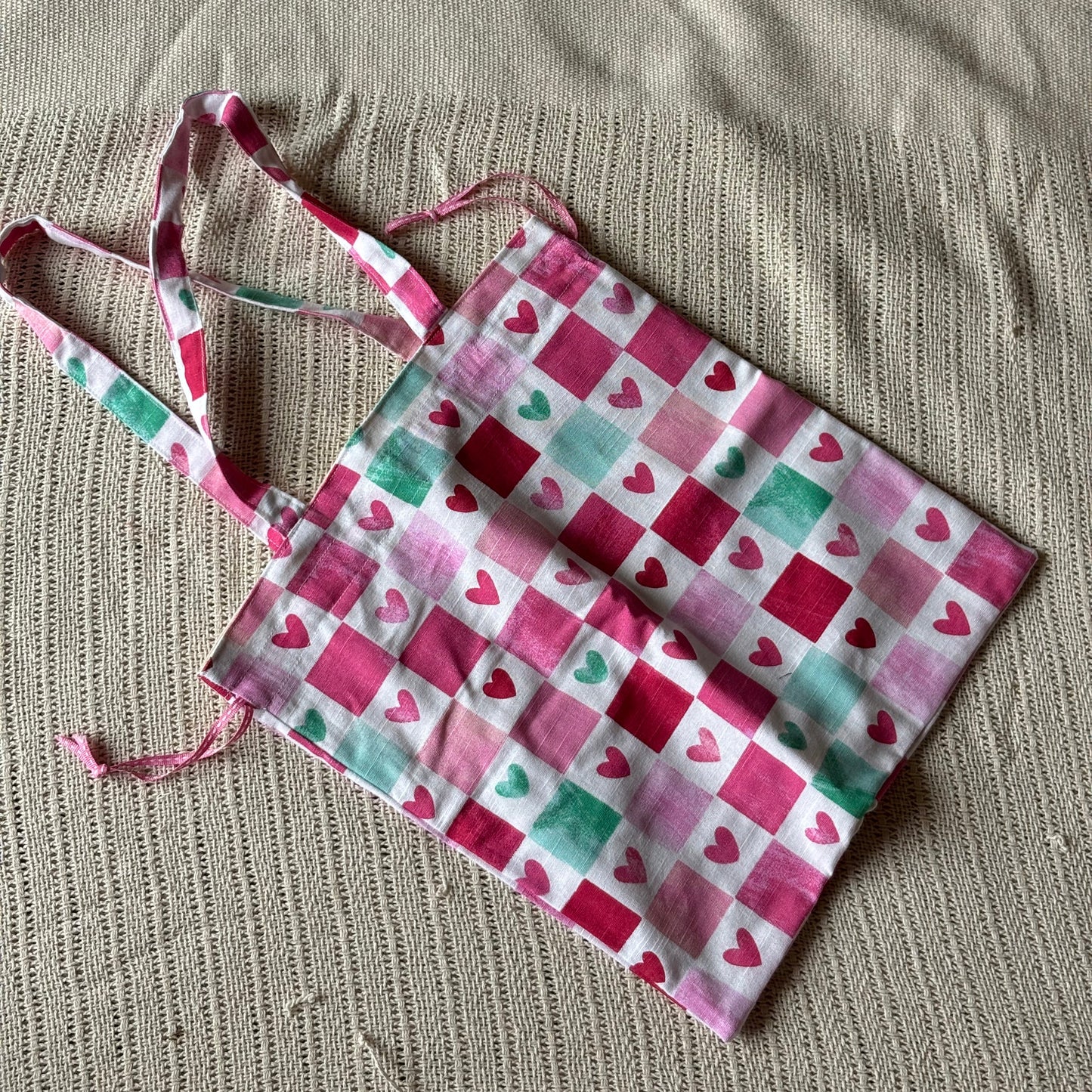 A fabric tote bag with a pink, red, and green checkered heart pattern, laid flat on a textured beige woven mat.