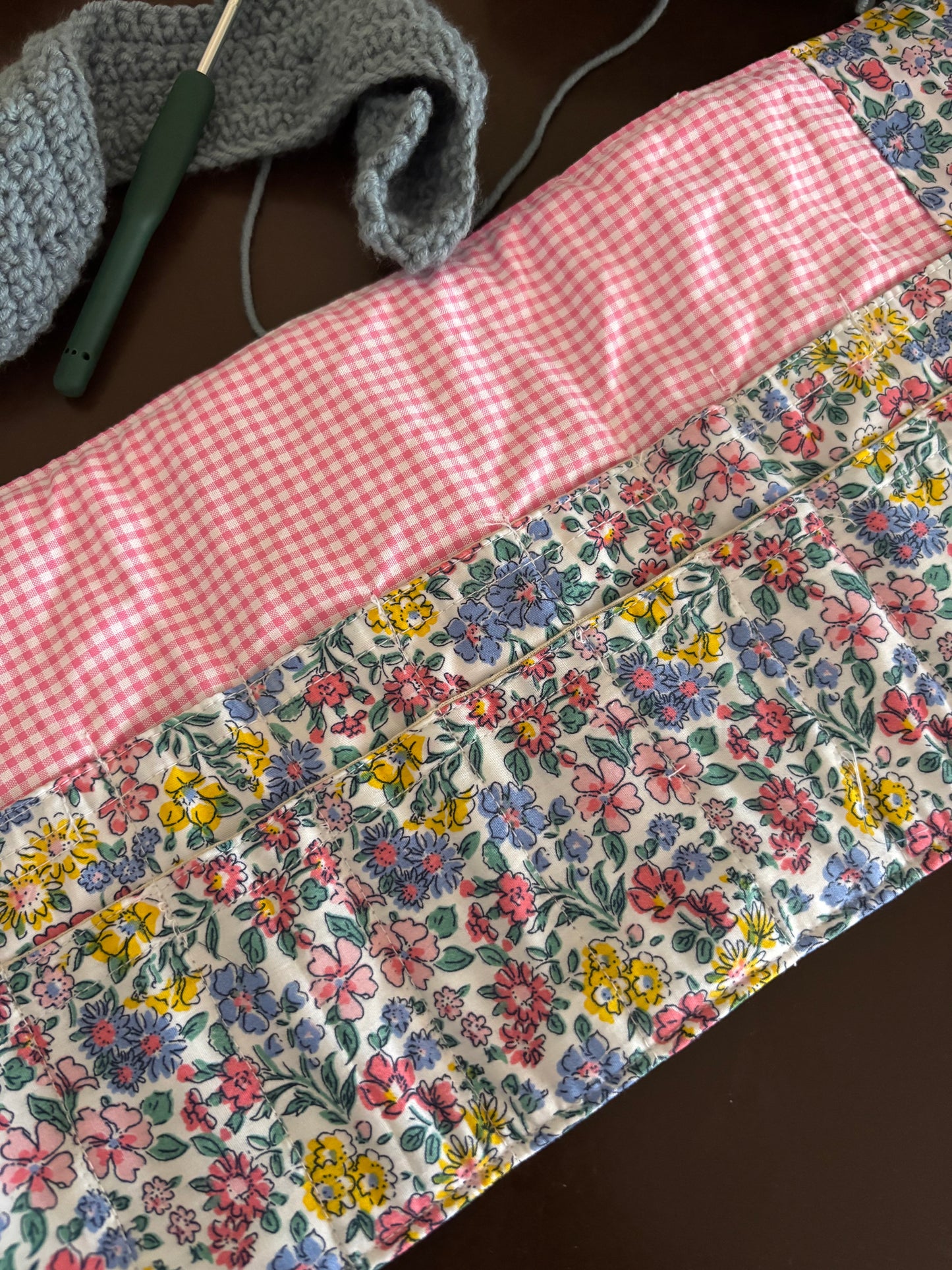 A handmade quilted fabric panel with a pink gingham checkered border and a colorful floral print center, shown on a dark brown surface with a crochet hook and yarn in the background.