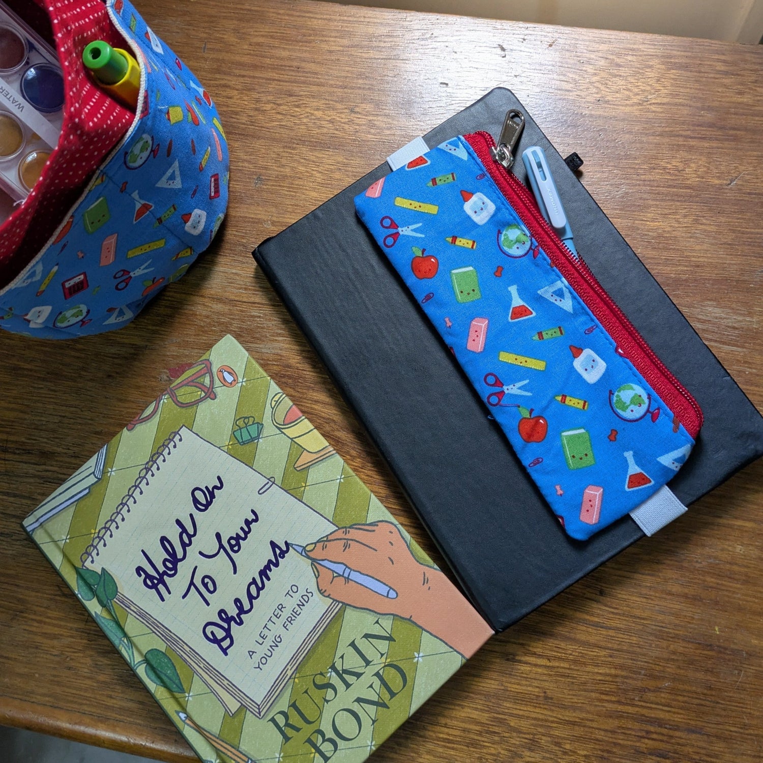 A blue pen sleeve with a pattern of books, pens, and other stationery items, displayed on top of a black hardbound journal or notepad.