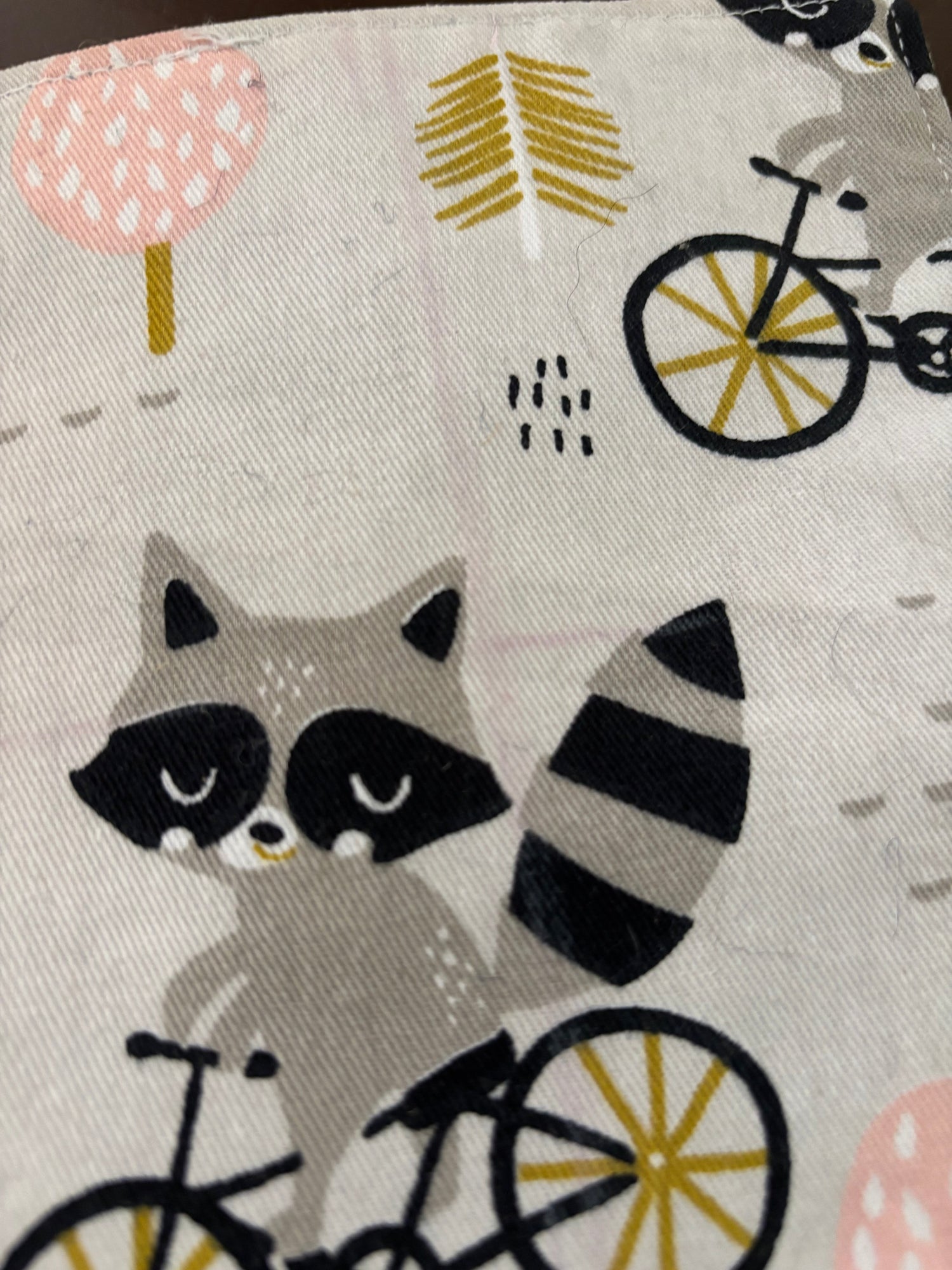 Close-up of a fabric surface with a whimsical raccoon and bicycle print on a light beige background. The design includes gray and black raccoons, black bicycles with yellow wheels, pink polka-dot shapes, and small yellow leaf motifs.