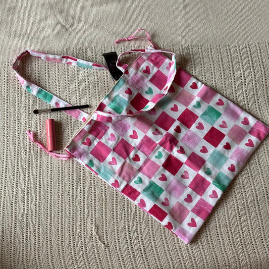 A handmade fabric tote bag with a pink, red, and mint green checkered heart pattern, laid flat on a beige textured knit blanket. A small pink lip balm tube and a black hairpin are placed next to the bag.