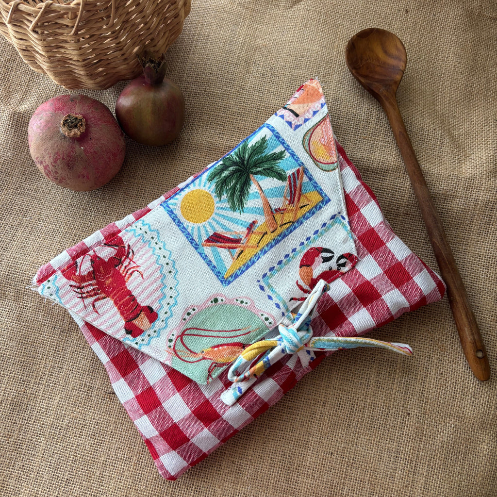 A fabric pouch with a red and white gingham base and a colorful patchwork-style top flap featuring lobster, beach, and sea creature motifs, tied with a braided cord. It is styled on a burlap surface with a wooden spoon, pomegranates, and a wicker basket nearby.
