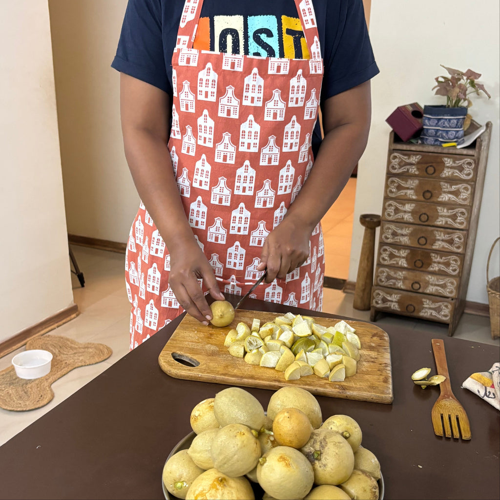 A woman wearing a navy blue t-shirt and a red apron with a white house pattern is slicing a potato on a wooden cutting board. In the foreground, a metal plate holds several unpeeled yellow melons on a dark brown table in a home kitchen setting.