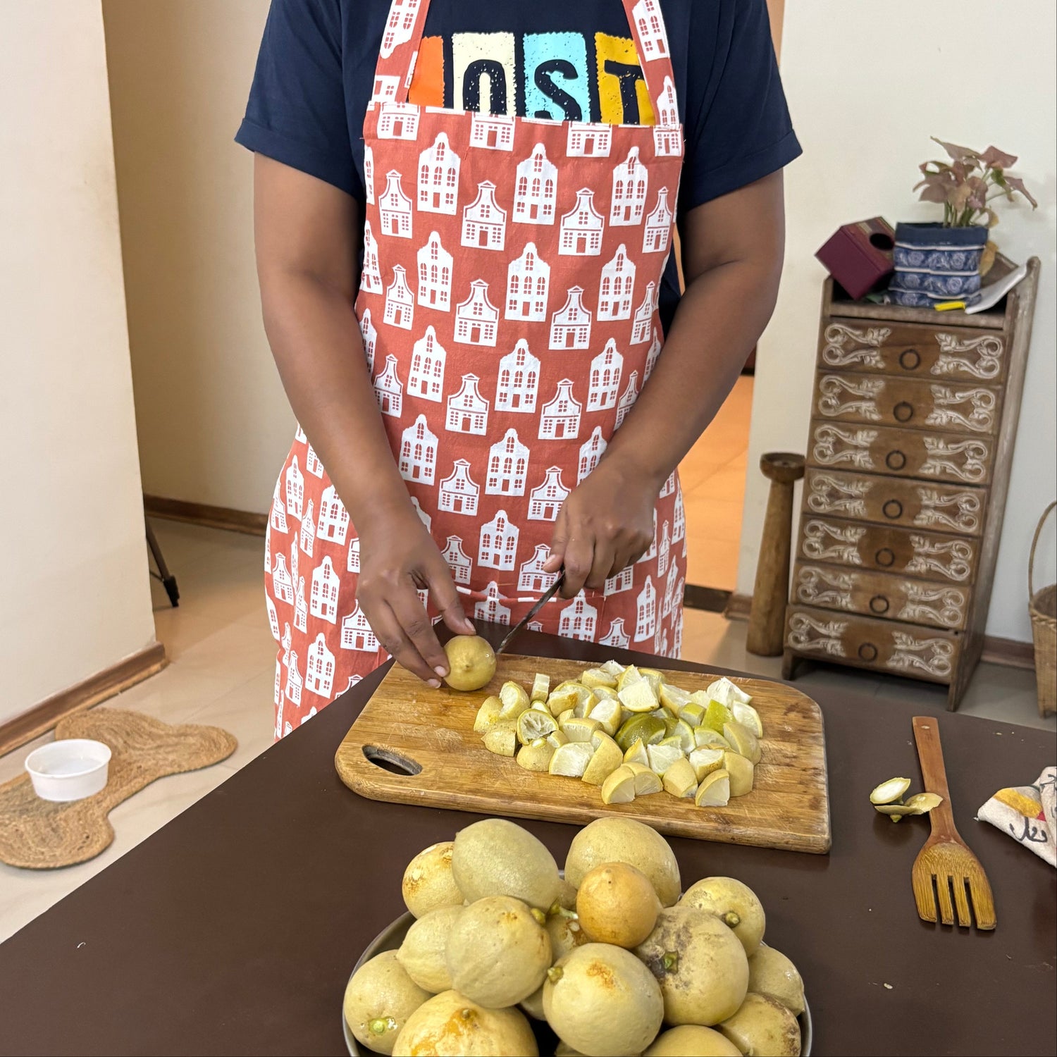 A woman wearing a navy blue t-shirt and a red apron with a white house pattern is slicing a potato on a wooden cutting board. In the foreground, a metal plate holds several unpeeled yellow melons on a dark brown table in a home kitchen setting.