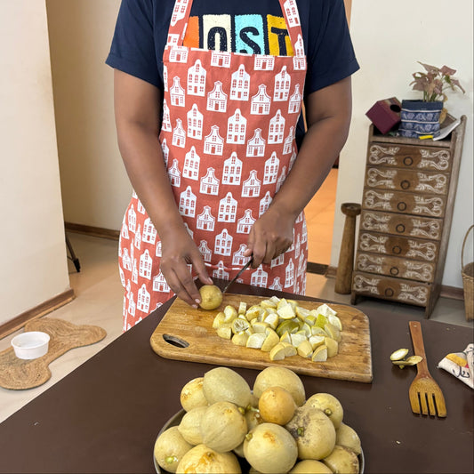 A woman wearing a navy blue t-shirt and a red apron with a white house pattern is slicing a potato on a wooden cutting board. In the foreground, a metal plate holds several unpeeled yellow melons on a dark brown table in a home kitchen setting.