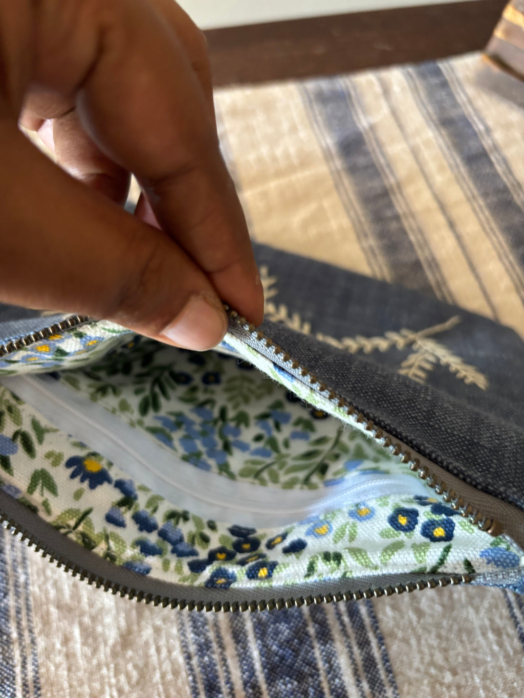Hand holding a floral-patterned fabric with a zipper, on a striped fabric background.