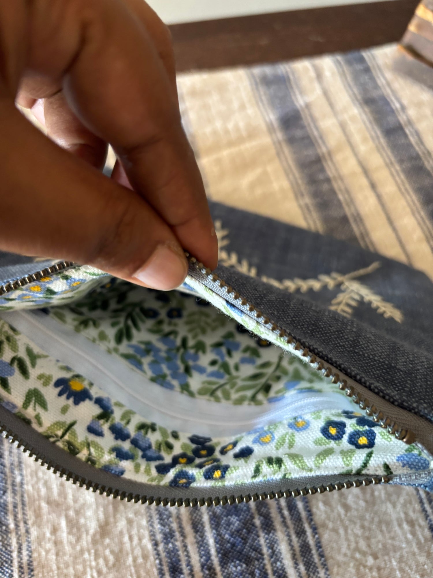 Hand holding a floral-patterned fabric with a zipper, on a striped fabric background.