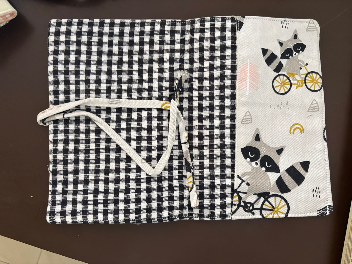 A fabric pouch or organizer cover laid flat on a dark brown surface, featuring a black-and-white gingham pattern on the right side and a white side panel with raccoon illustrations and bicycle motifs. A white drawstring is attached near the center.