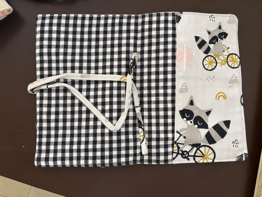 A fabric pouch or organizer cover laid flat on a dark brown surface, featuring a black-and-white gingham pattern on the right side and a white side panel with raccoon illustrations and bicycle motifs. A white drawstring is attached near the center.