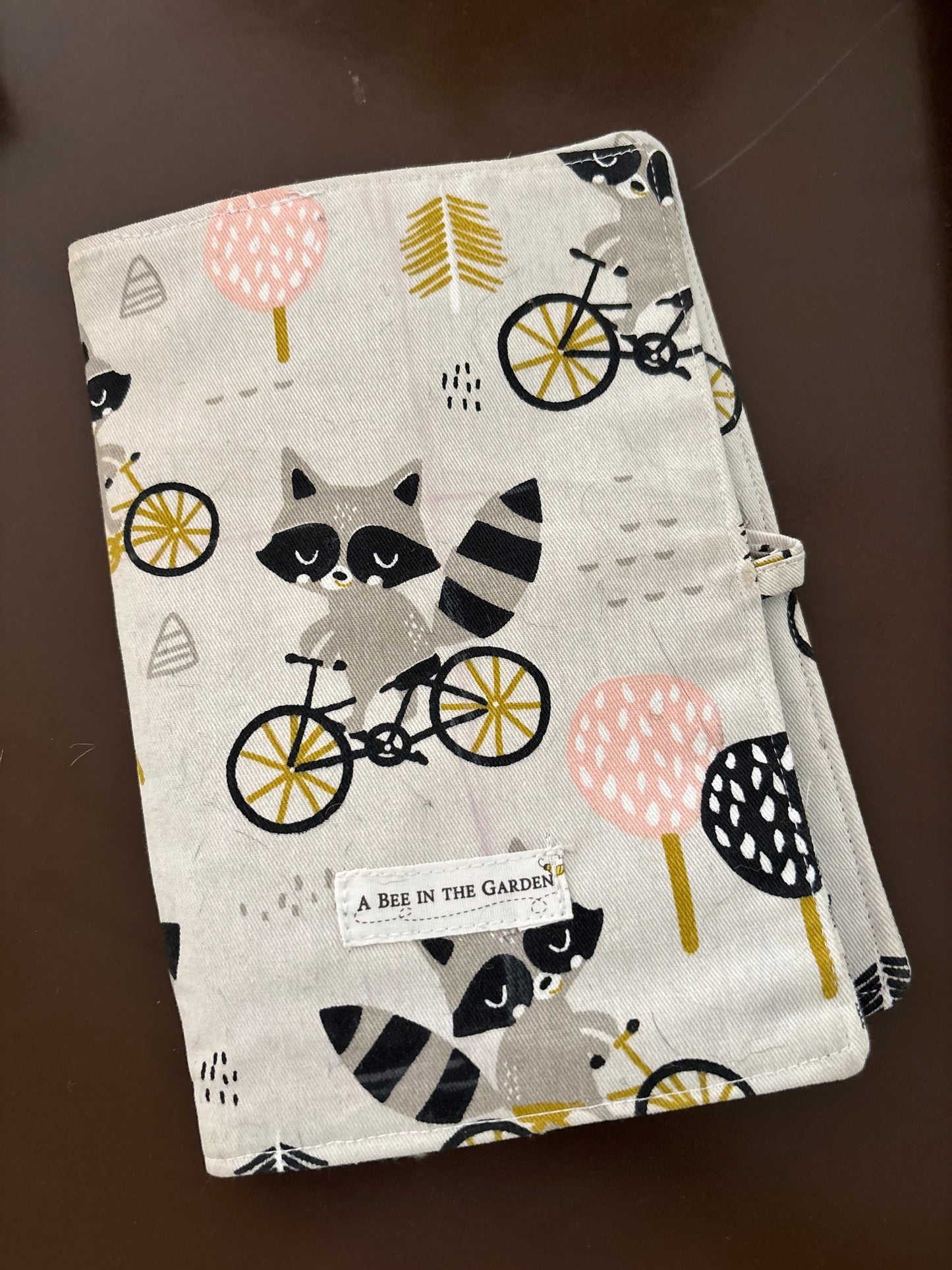 A fabric zippered pouch or clutch with a whimsical raccoon and bicycle print, shown on a dark brown surface. The print features gray and black raccoons on bicycles with pink, yellow, and black accents.
