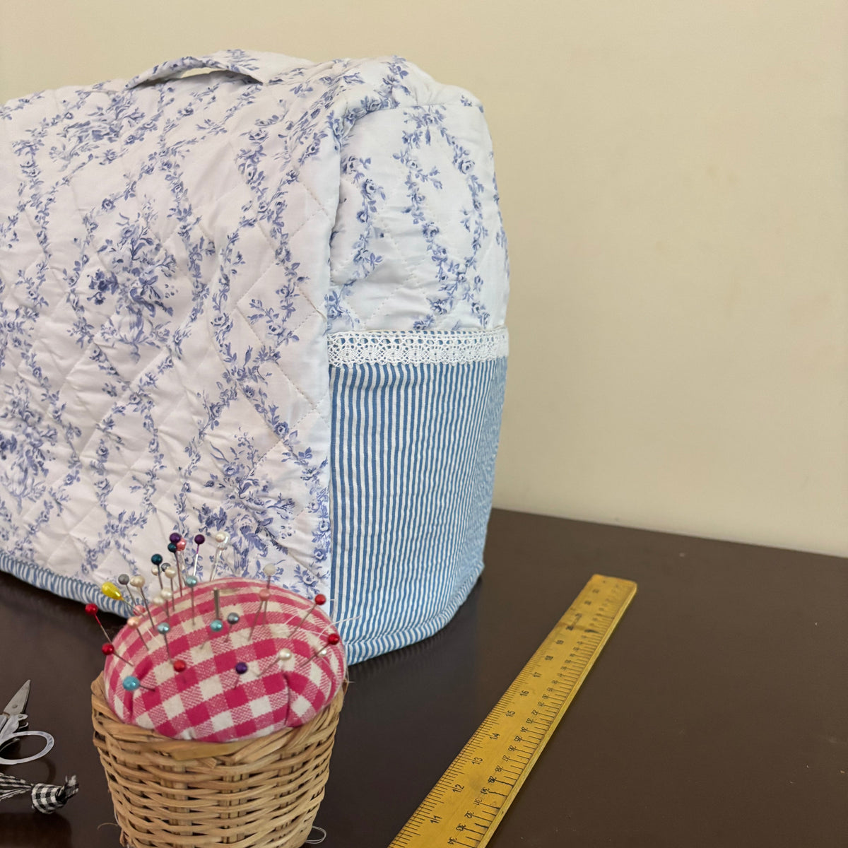 A handmade sewing machine cover with a blue and white floral pattern and a striped side pocket, shown on a dark table next to a yellow measuring tape and a small basket with a gingham pincushion.