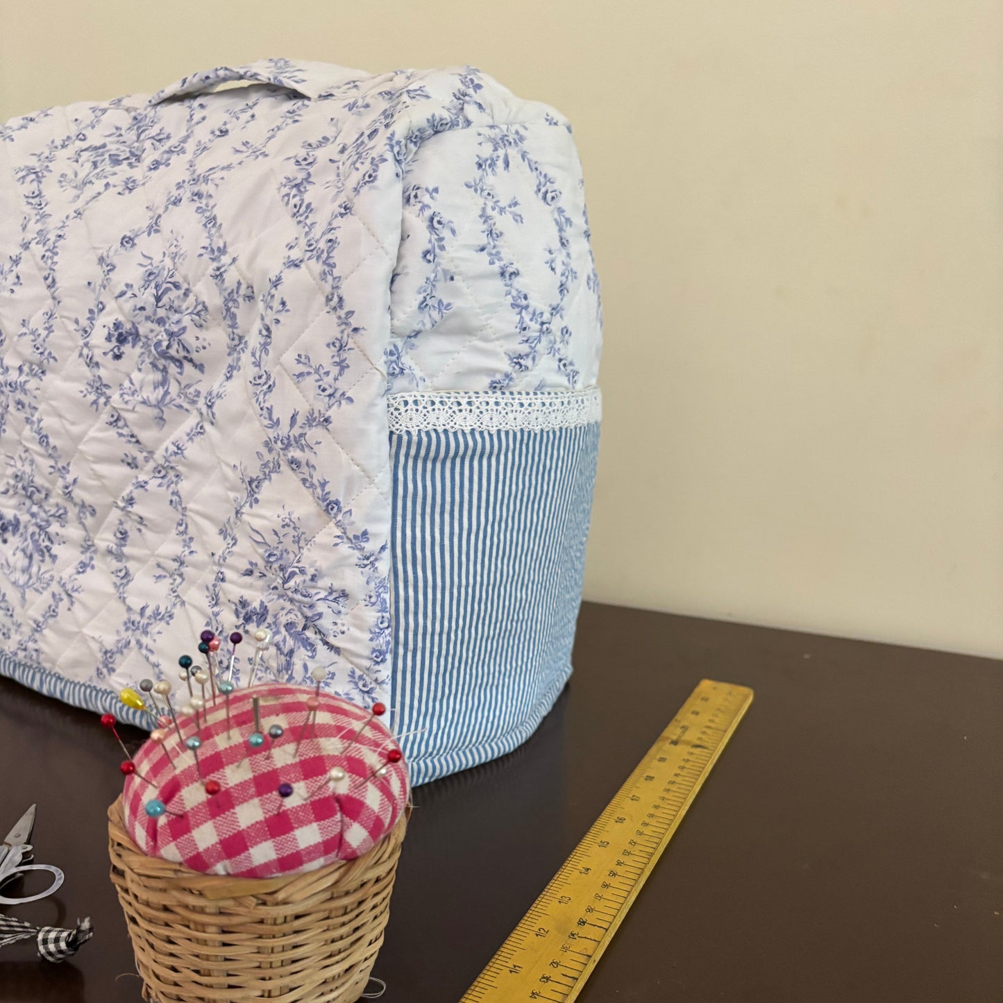 A handmade sewing machine cover with a blue and white floral pattern and a striped side pocket, shown on a dark table next to a yellow measuring tape and a small basket with a gingham pincushion.