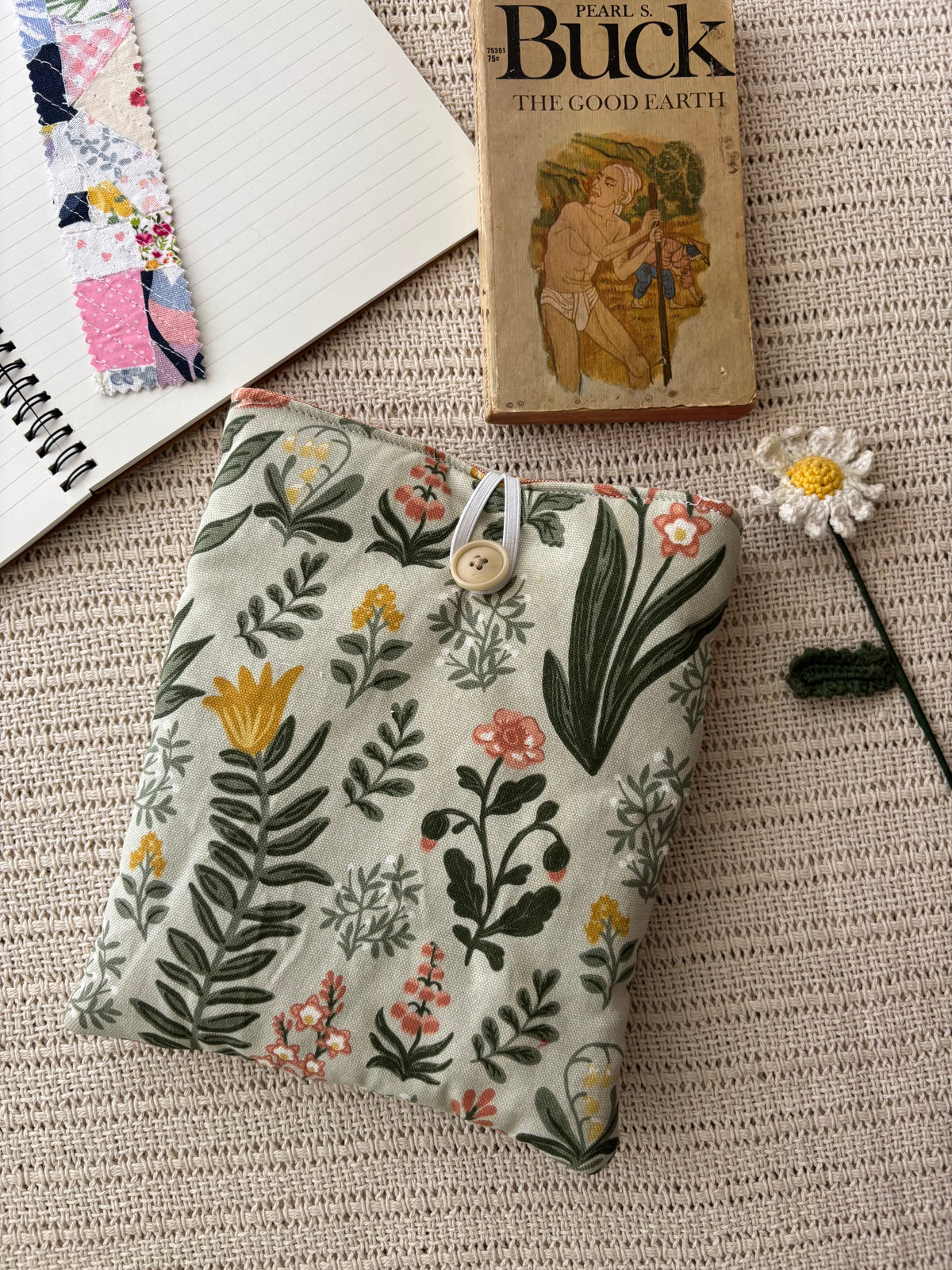 A fabric pouch with a botanical floral print in muted green, beige, yellow, and pink, styled on a textured beige woven blanket alongside a vintage Pearl Buck book, a notebook, and a small daisy.