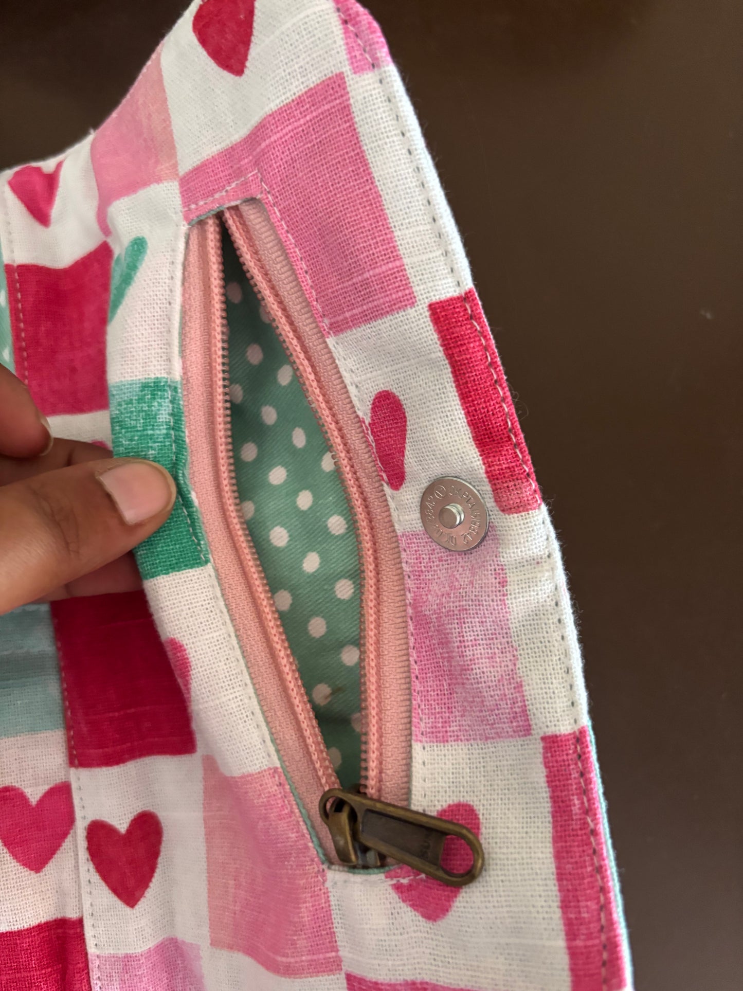 Close-up of a fabric pouch with a pink and white checkered heart pattern, a pink zipper, and a green polka-dot interior lining, held by a hand.