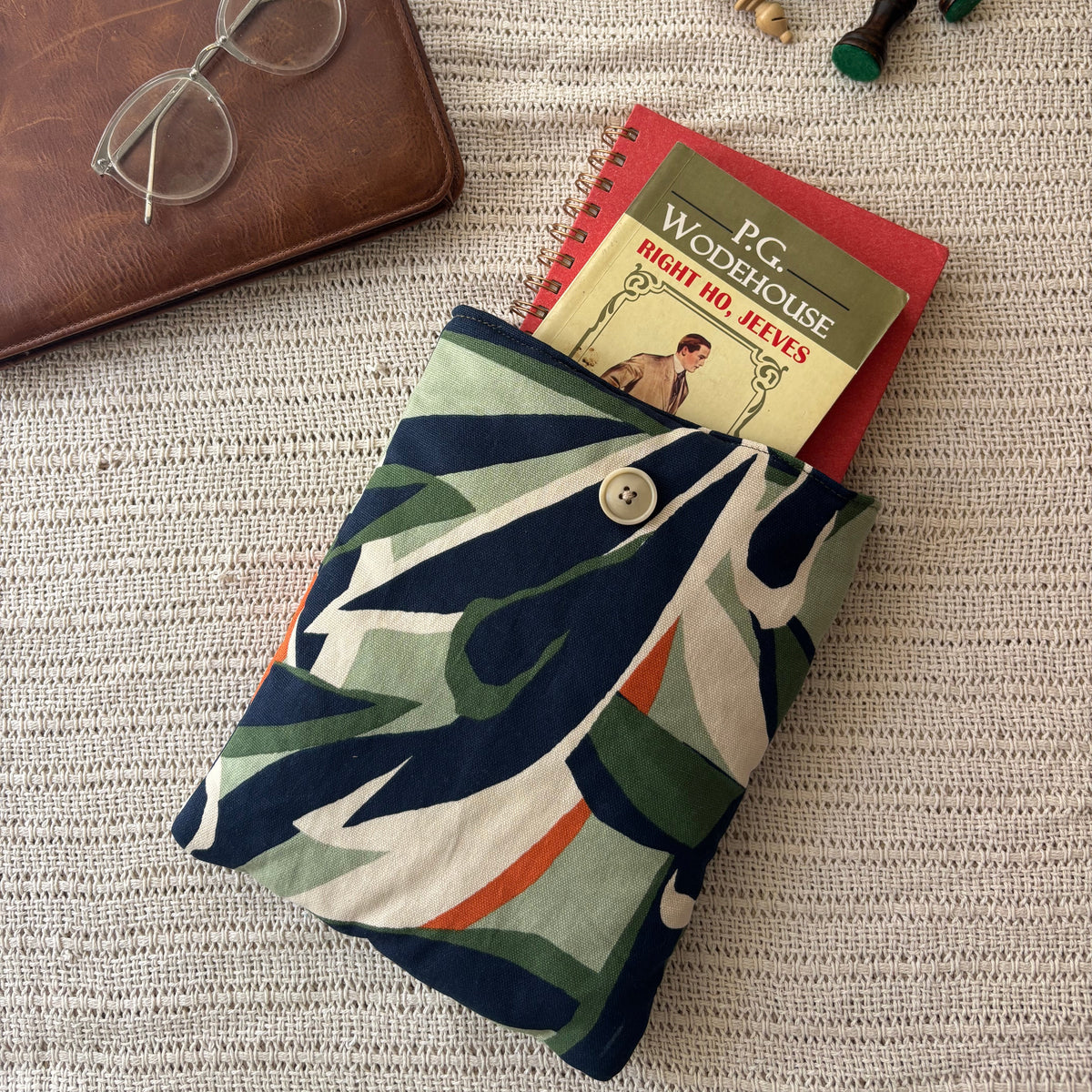 A fabric book pouch with a colorful abstract pattern in navy, green, orange, and cream, shown holding a red spiral notebook. The pouch is styled on a textured beige woven blanket alongside a brown leather notebook, clear eyeglasses, and wooden chess pieces.