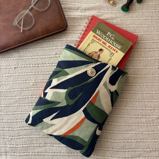 A fabric book pouch with a colorful abstract pattern in navy, green, orange, and cream, shown holding a red spiral notebook. The pouch is styled on a textured beige woven blanket alongside a brown leather notebook, clear eyeglasses, and wooden chess pieces.