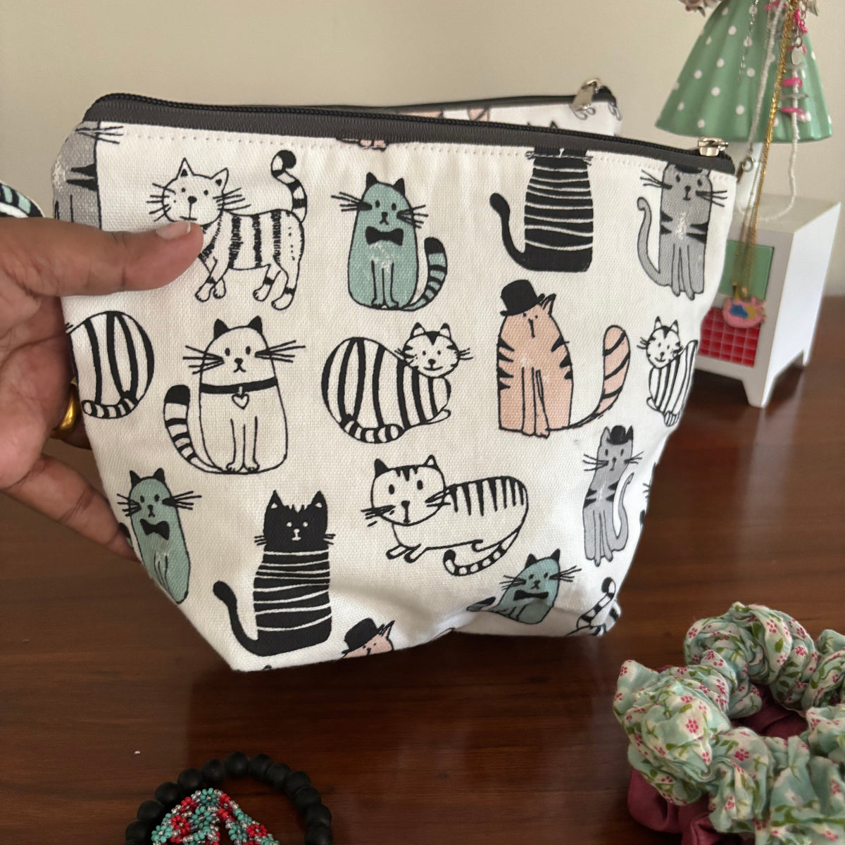 Small pouch with cat pattern held by a hand on a wooden surface with scrunchies and a bracelet.