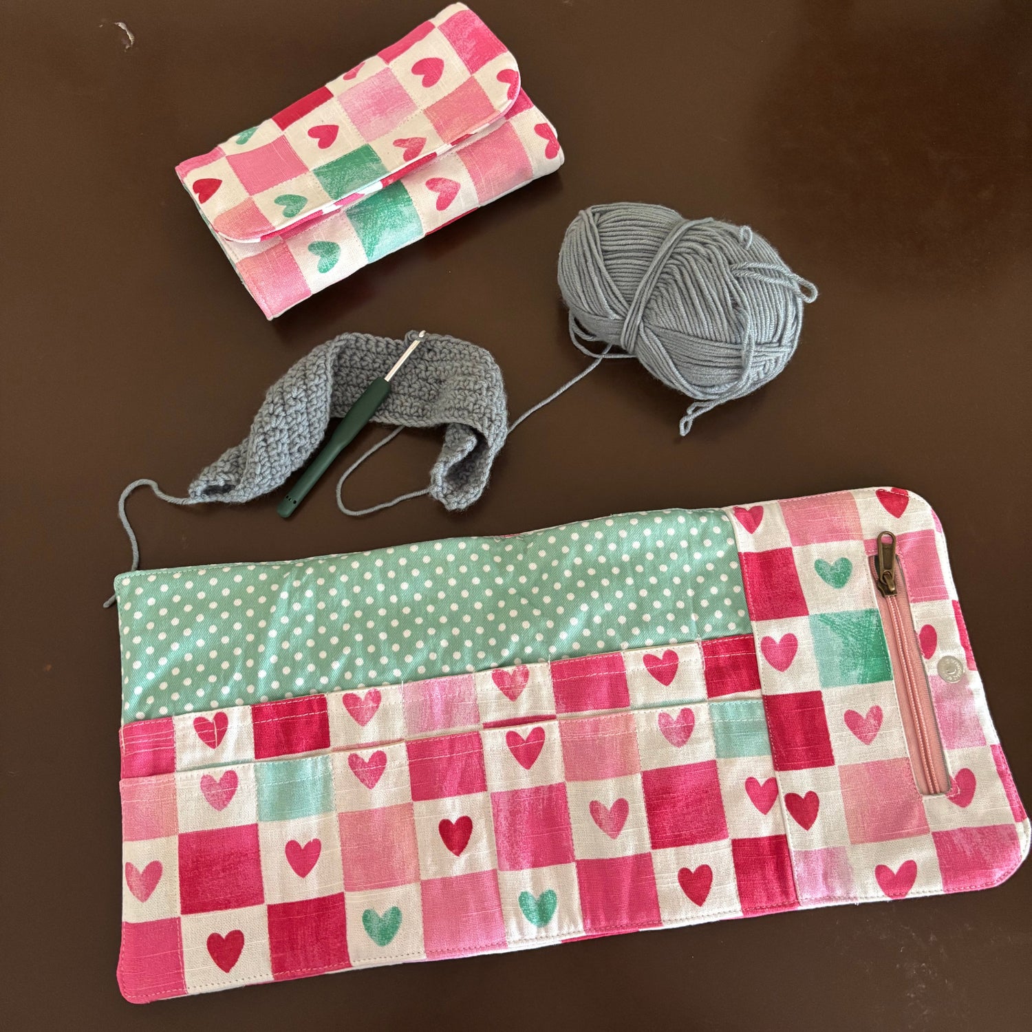 A handmade patchwork fabric pouch with pink and green heart patterns, shown alongside a gray crochet hook, gray yarn, and a smaller matching pouch on a dark brown surface.