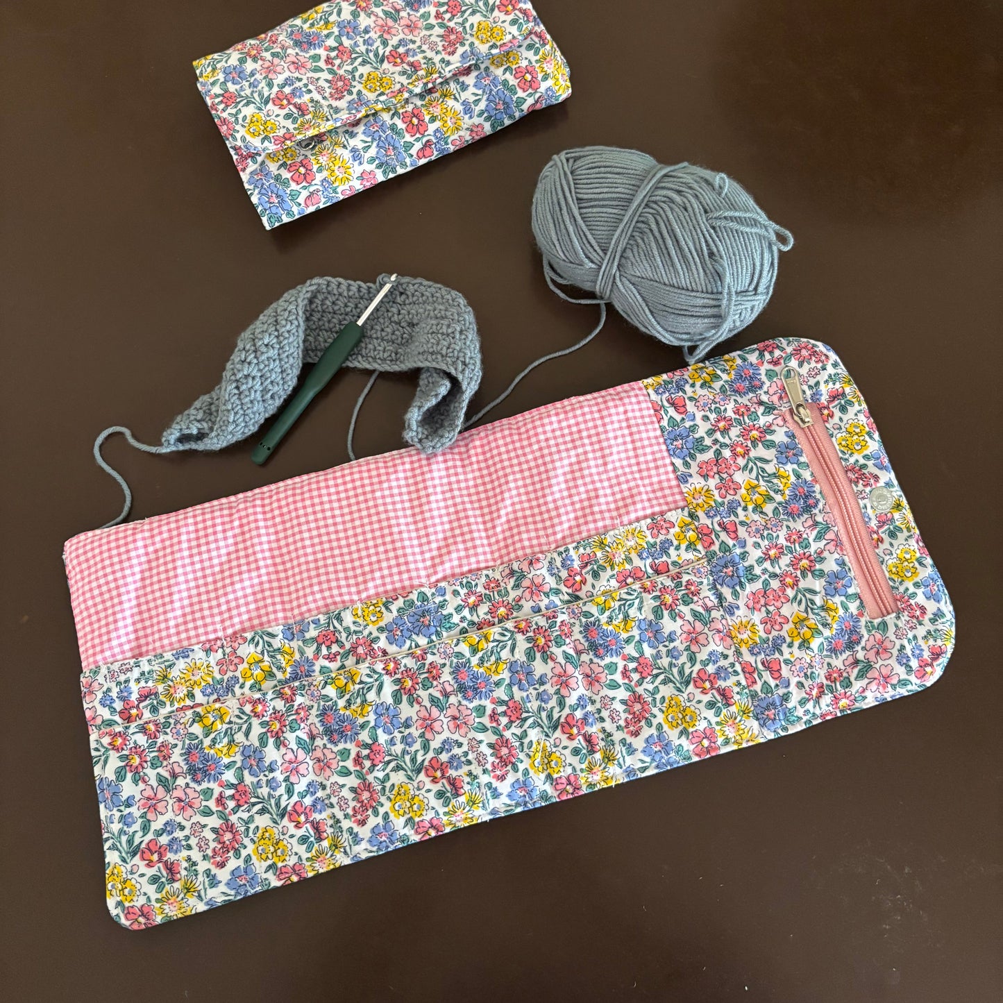 A handmade fabric pouch with a floral print and pink gingham panel, shown with a crochet hook, gray yarn, and a smaller matching pouch on a dark brown surface.