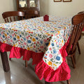 A rectangular dining table covered with a white tablecloth featuring a colorful floral and bird pattern, with red ruffled table skirt and matching red ruffled chair covers on wooden chairs in a dining room.