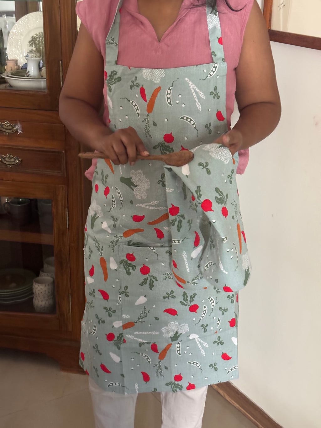 A person wearing a sage green apron with a colorful vegetable print (carrots, peas, radishes) and holding a matching cloth napkin in a home kitchen setting.