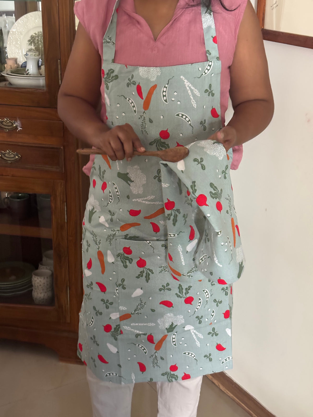 A person wearing a sage green apron with a colorful vegetable print (carrots, peas, radishes) and holding a matching cloth napkin in a home kitchen setting.