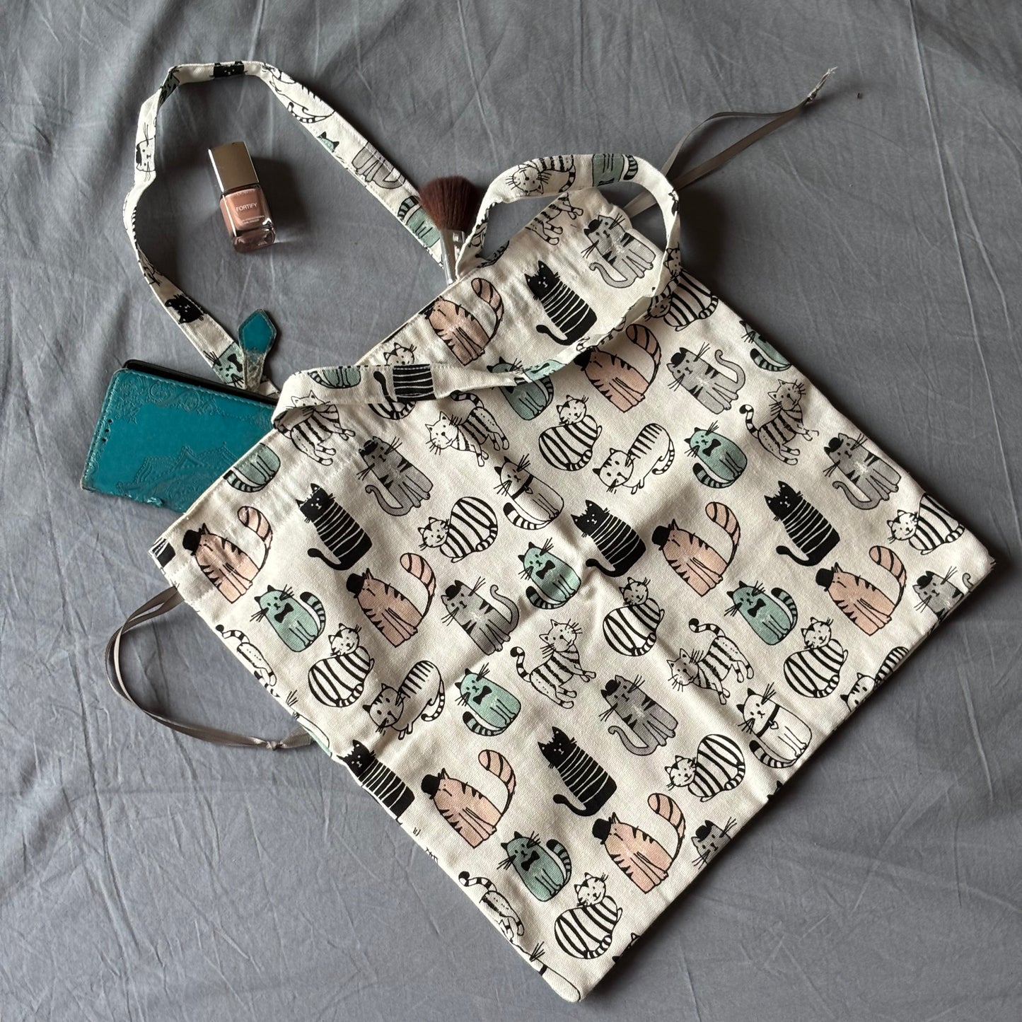 A fabric tote bag with a cream background and a repeating pattern of illustrated striped and cat-shaped bags, laid flat on a gray fabric surface with small accessories placed beside it.