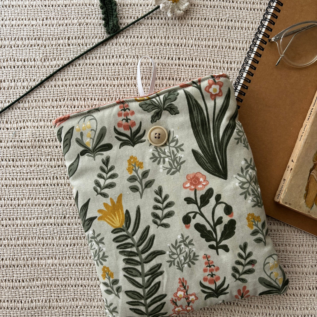 A fabric pouch with a botanical floral print in green, yellow, and pink on a light beige background, styled on a textured cream woven blanket with a spiral notebook, glasses, and a crochet flower.