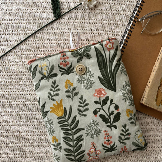 A fabric pouch with a botanical floral print in green, yellow, and pink on a light beige background, styled on a textured cream woven blanket with a spiral notebook, glasses, and a crochet flower.