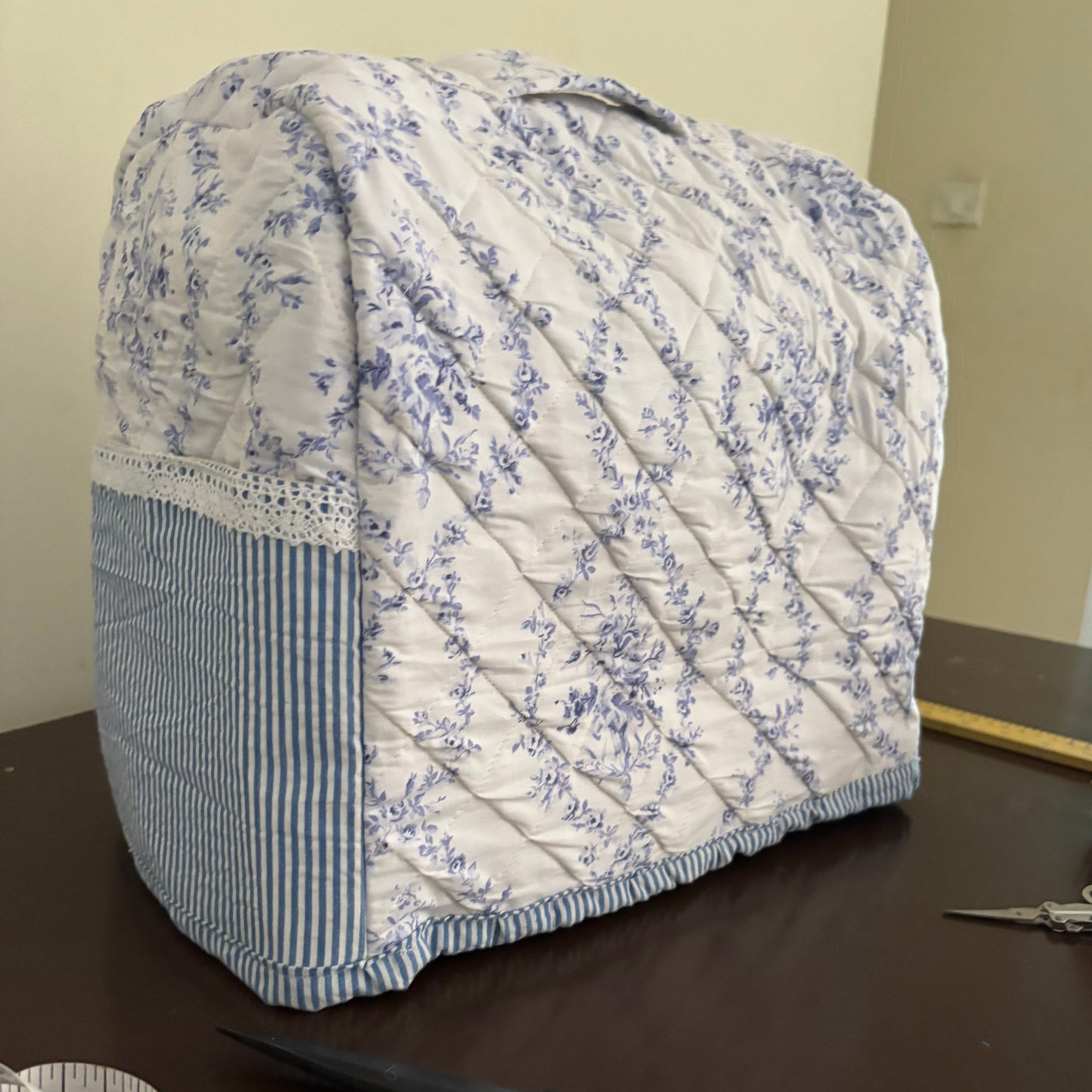 A quilted sewing machine cover with a blue floral pattern on a white background and a striped side panel, shown on a dark wooden table with scissors and a measuring tape in the foreground.