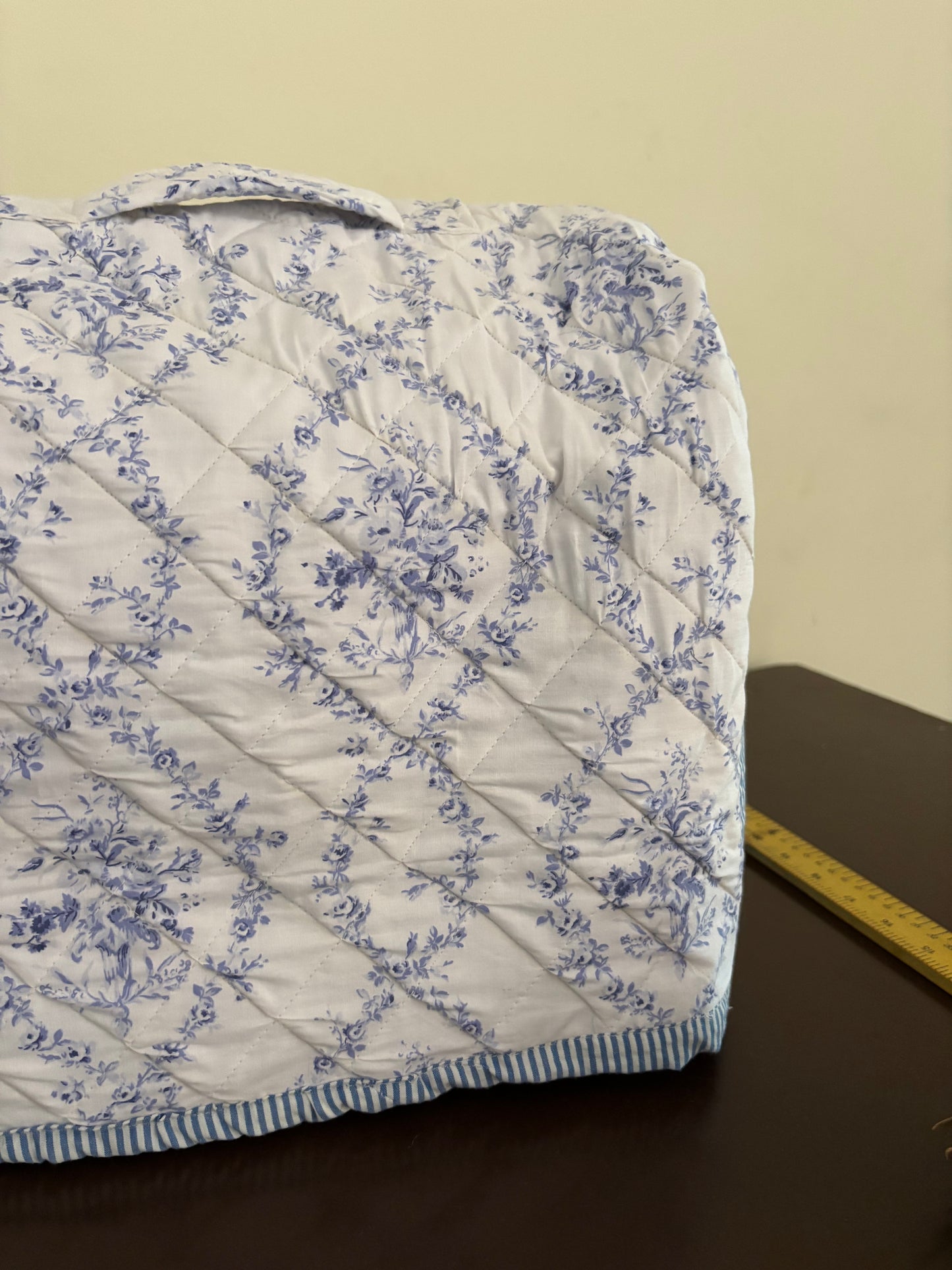 A quilted fabric cover with a blue floral toile pattern on a white background, shown on a dark table with a yellow measuring tape visible on the right side.