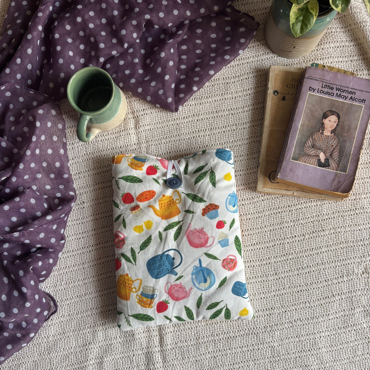A folded quilted blanket with a colorful teapot and floral pattern on a white background, styled on a textured beige blanket alongside a purple polka-dot throw, a green mug, a book, and a potted plant.
