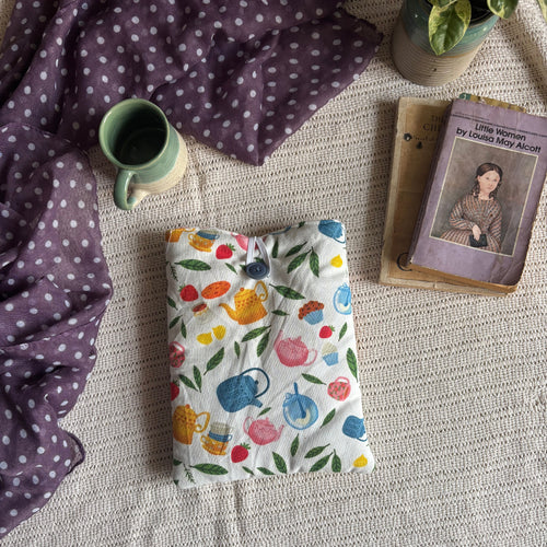 A folded quilted blanket with a colorful teapot and floral pattern on a white background, styled on a textured beige blanket alongside a purple polka-dot throw, a green mug, a book, and a potted plant.