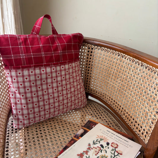 Red plaid bag and pillow on a wicker chair with a book featuring floral illustrations.