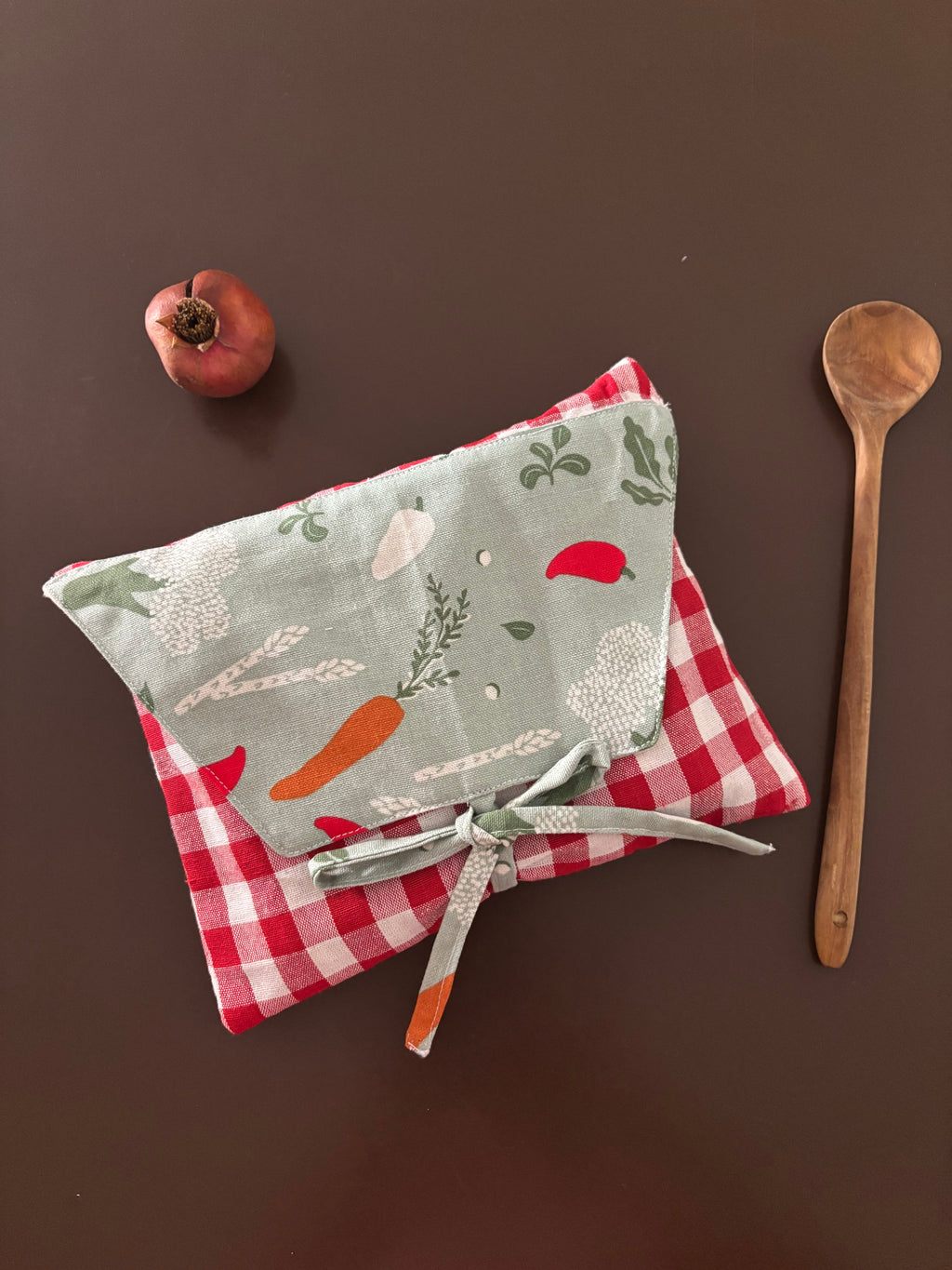 A fabric kitchen towel or apron-style cloth with a red-and-white gingham base and a pale sage green front panel printed with vegetables, tied with a matching sage ribbon. It is photographed on a dark brown surface with a wooden spoon and a small red fruit nearby.