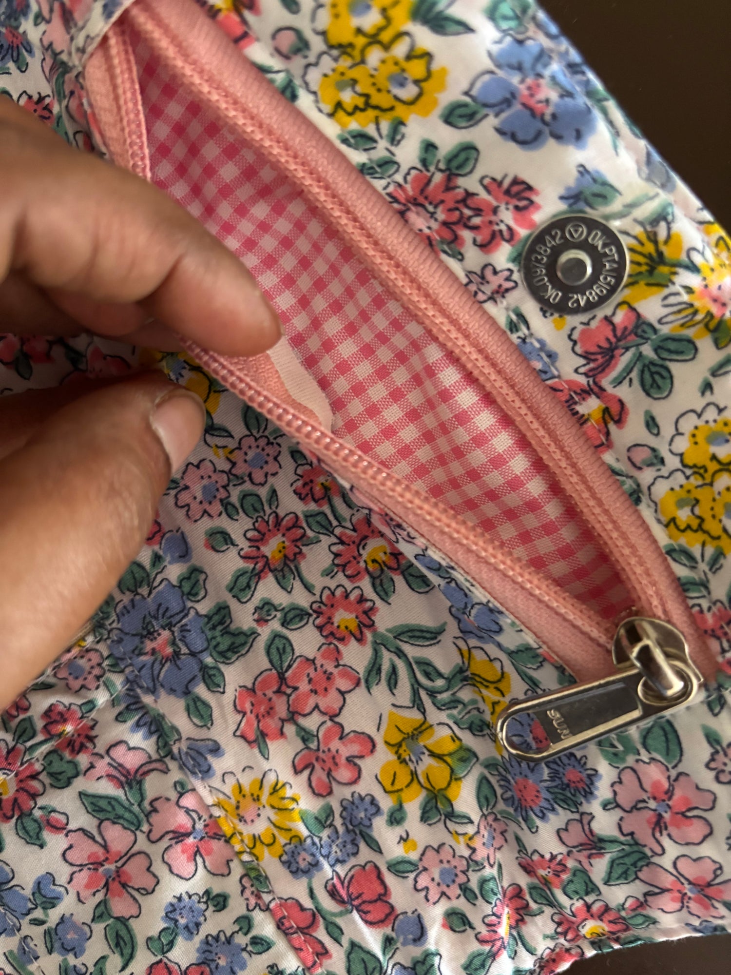Close-up of a floral zippered pouch with a hand opening the zipper to show a pink gingham lining. The pouch has a white background with a multicolor floral print in pink, yellow, blue, and green, and a silver metal snap button closure.