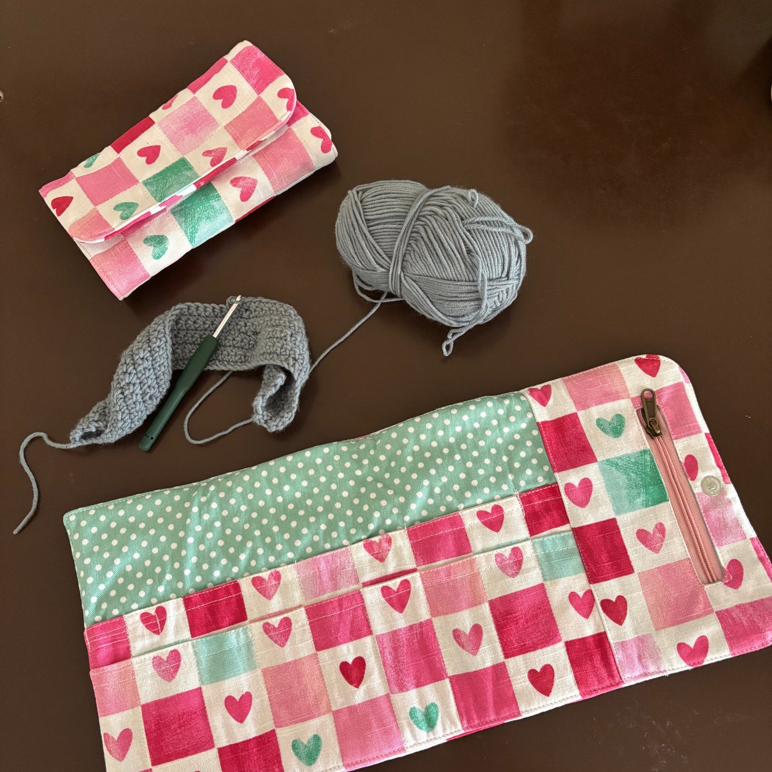 A handmade patchwork pouch with pink, red, and mint green heart patterns, shown alongside a matching zippered pouch, gray yarn, and a crochet hook on a dark brown wooden surface.