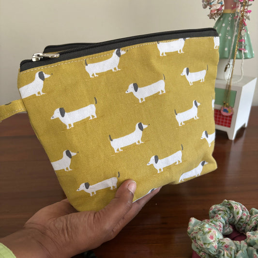 Yellow pouch with dog pattern held by a hand, with a wooden surface and decorative items in the background.