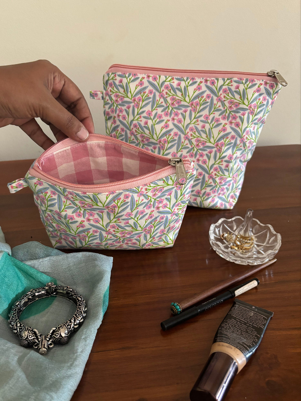 Hand holding a floral-patterned makeup bag with cosmetic items on a wooden surface.