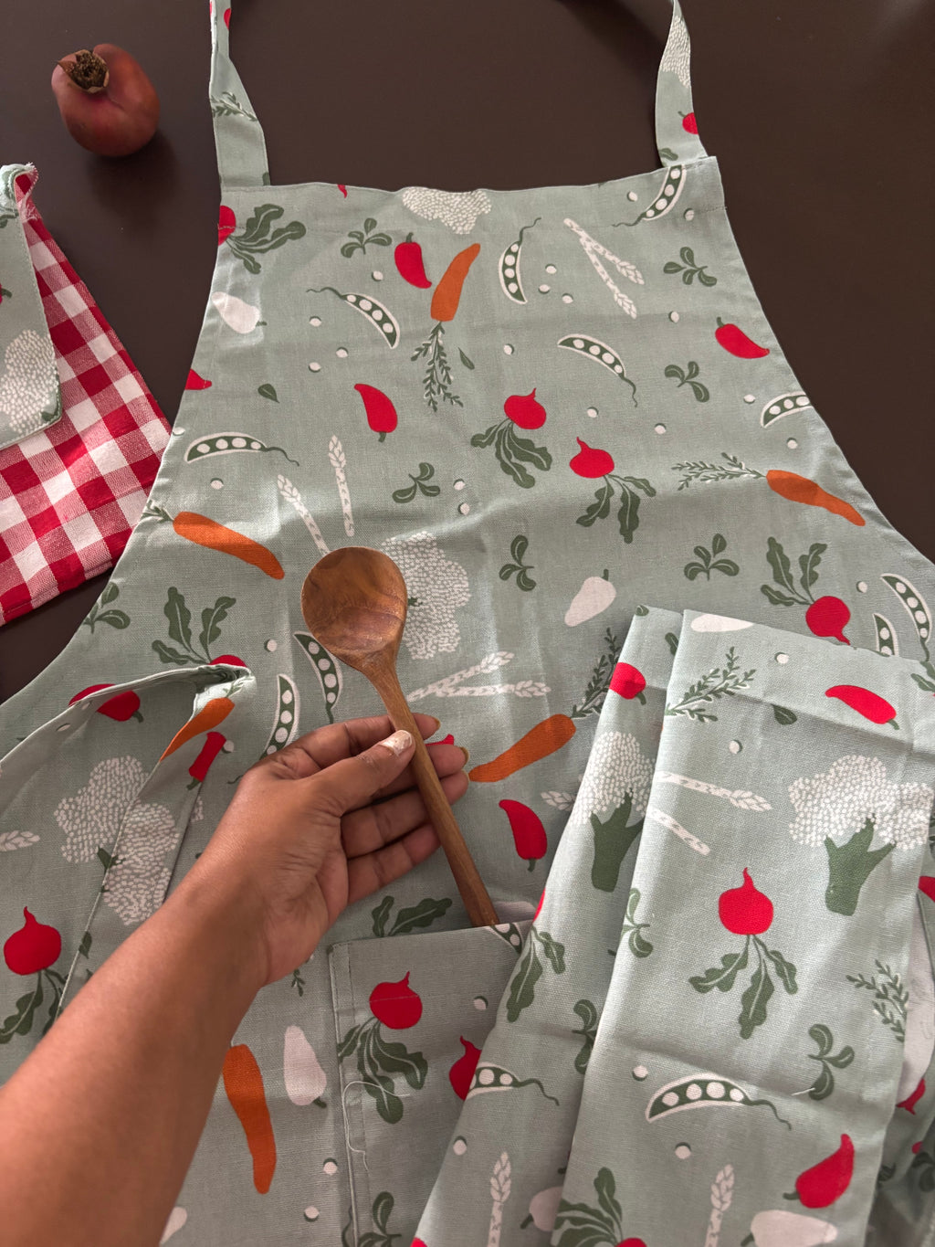 A sage green apron with a vegetable print featuring red radishes, orange carrots, and green leaves, shown with a person's hand holding a wooden spoon in the front pocket.