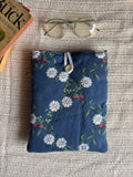 A navy blue fabric pouch with a white daisy and green leaf floral pattern, styled on a beige woven blanket with a pair of glasses and a book corner visible.