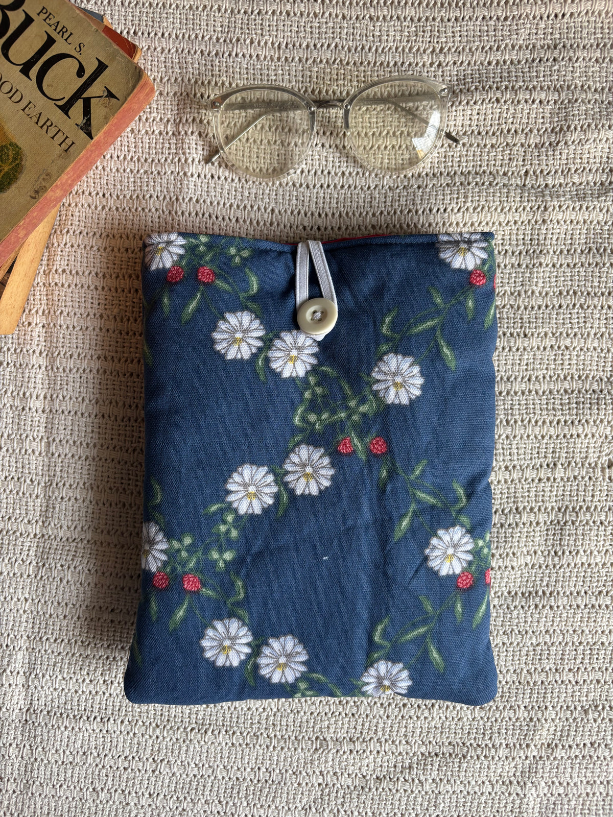 A navy blue fabric pouch with a white daisy and green leaf floral pattern, styled on a beige woven blanket with a pair of glasses and a book corner visible.