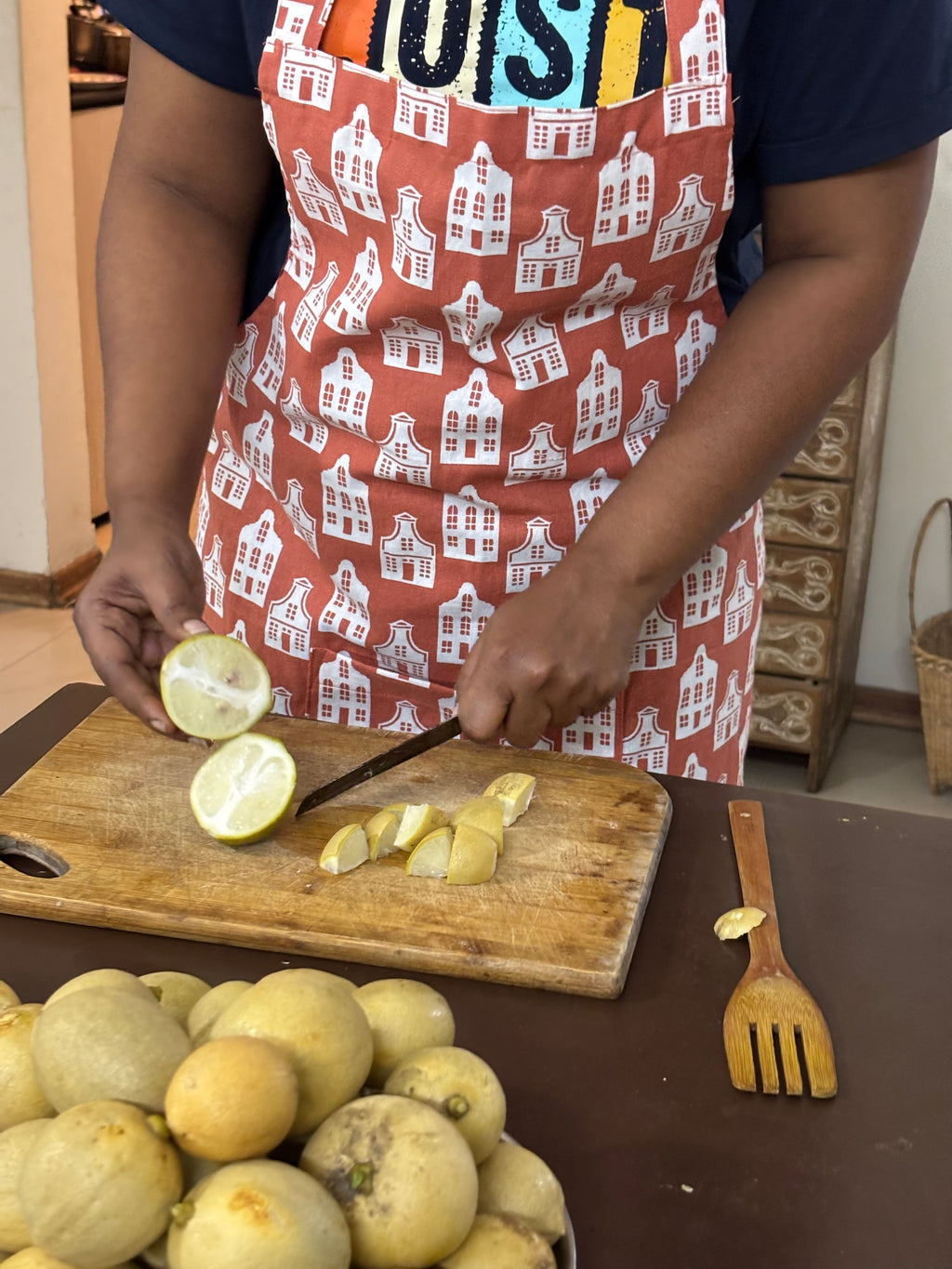 A person wearing a rust-orange apron with a white house patternc, slicing yellow citrus fruit on a wooden cutting board in a kitchen setting.