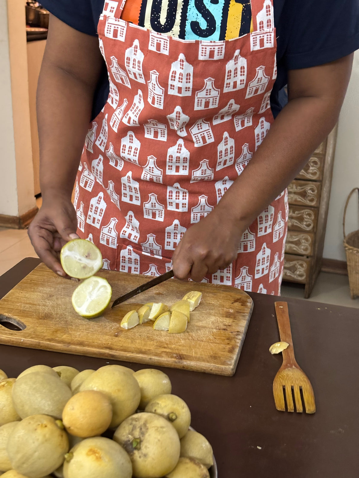 A person wearing a rust-orange apron with a white house patternc, slicing yellow citrus fruit on a wooden cutting board in a kitchen setting.