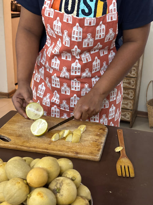 A person wearing a rust-orange apron with a white house patternc, slicing yellow citrus fruit on a wooden cutting board in a kitchen setting.