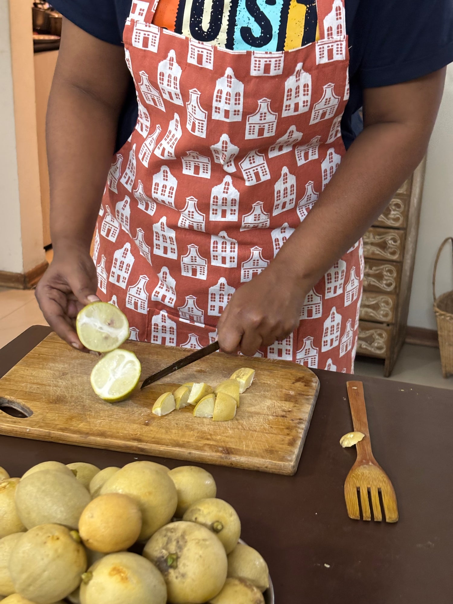 A person wearing a rust-orange apron with a white house patternc, slicing yellow citrus fruit on a wooden cutting board in a kitchen setting.