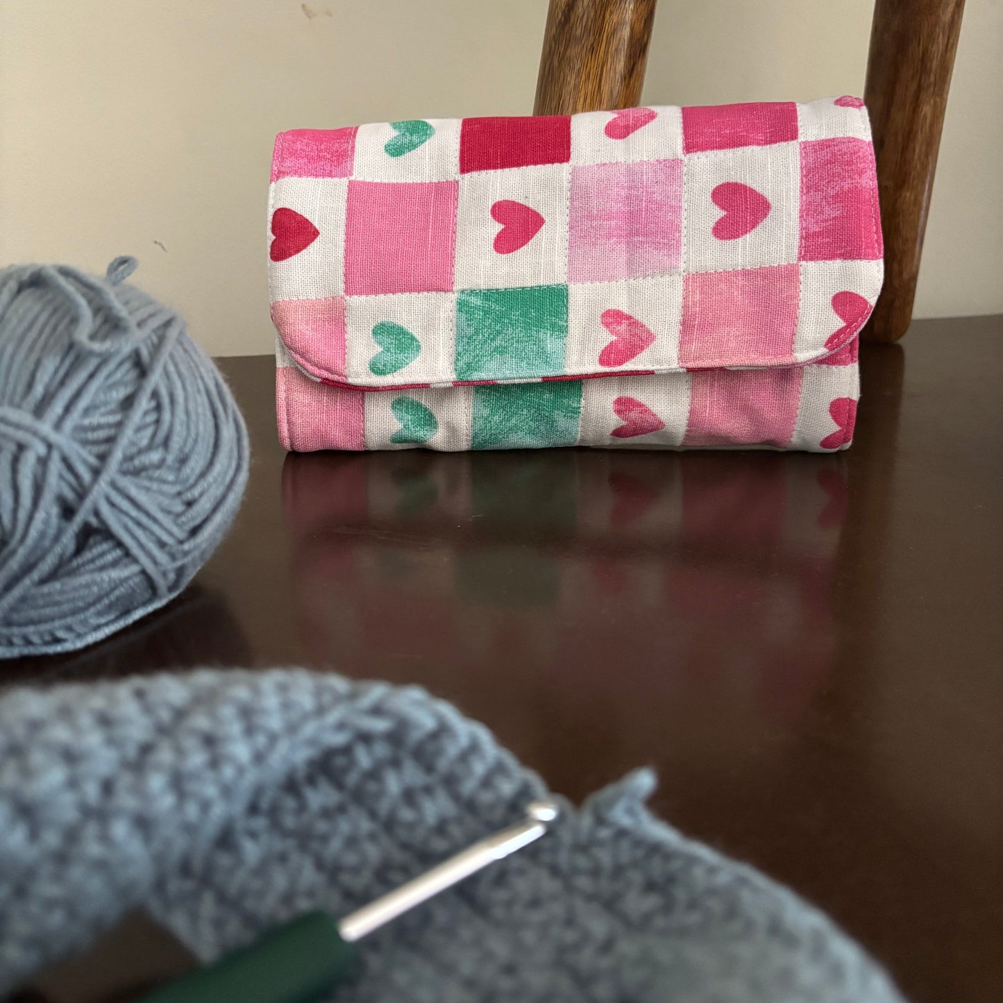 A handmade fabric clutch or pouch with a pink, red, and green heart pattern on a white background, resting on a dark wooden table next to a ball of gray yarn and a crochet hook.