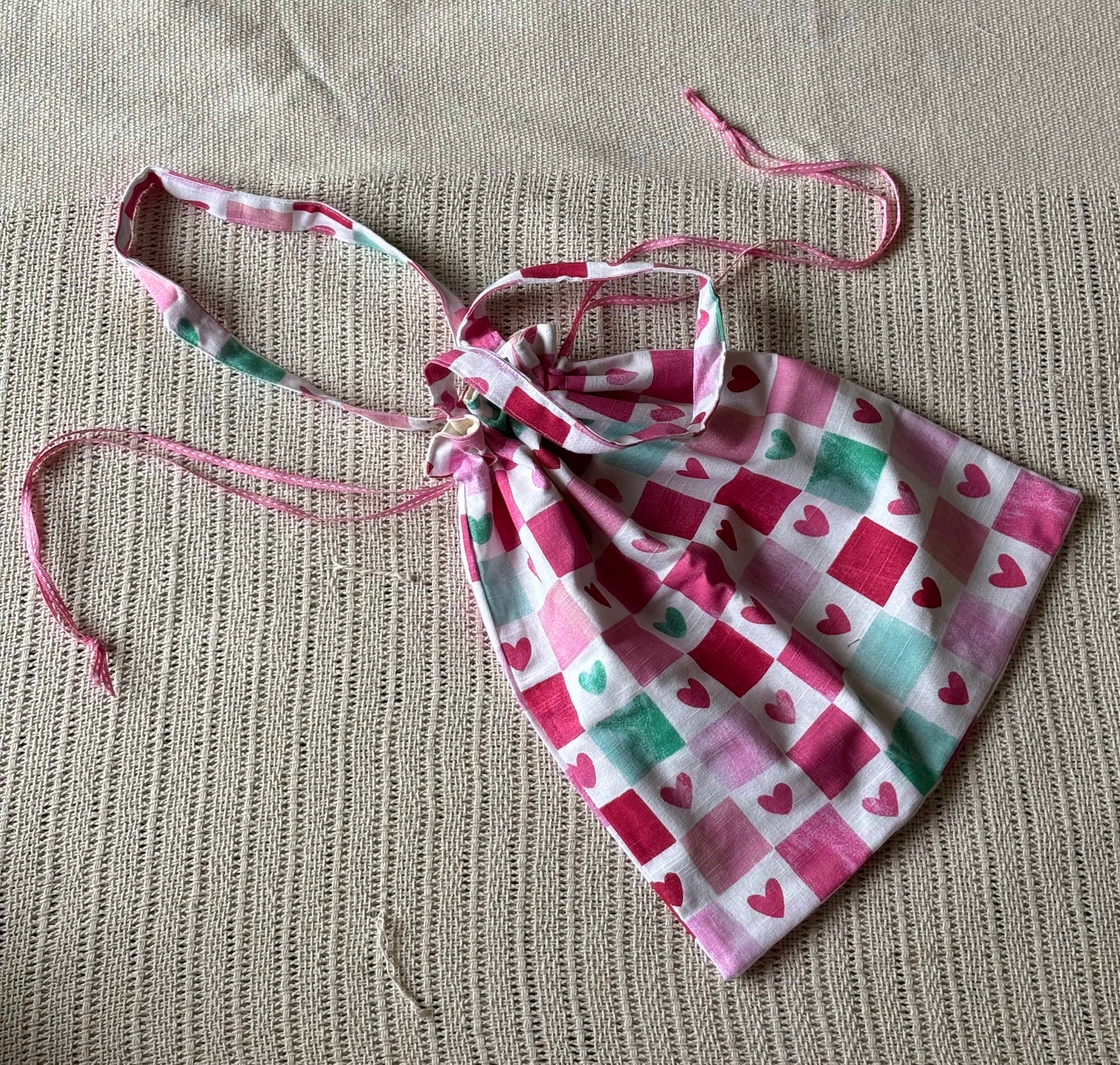 A small fabric drawstring pouch with a pink, white, and mint green checkered pattern featuring hearts, laid flat on a textured beige woven mat.