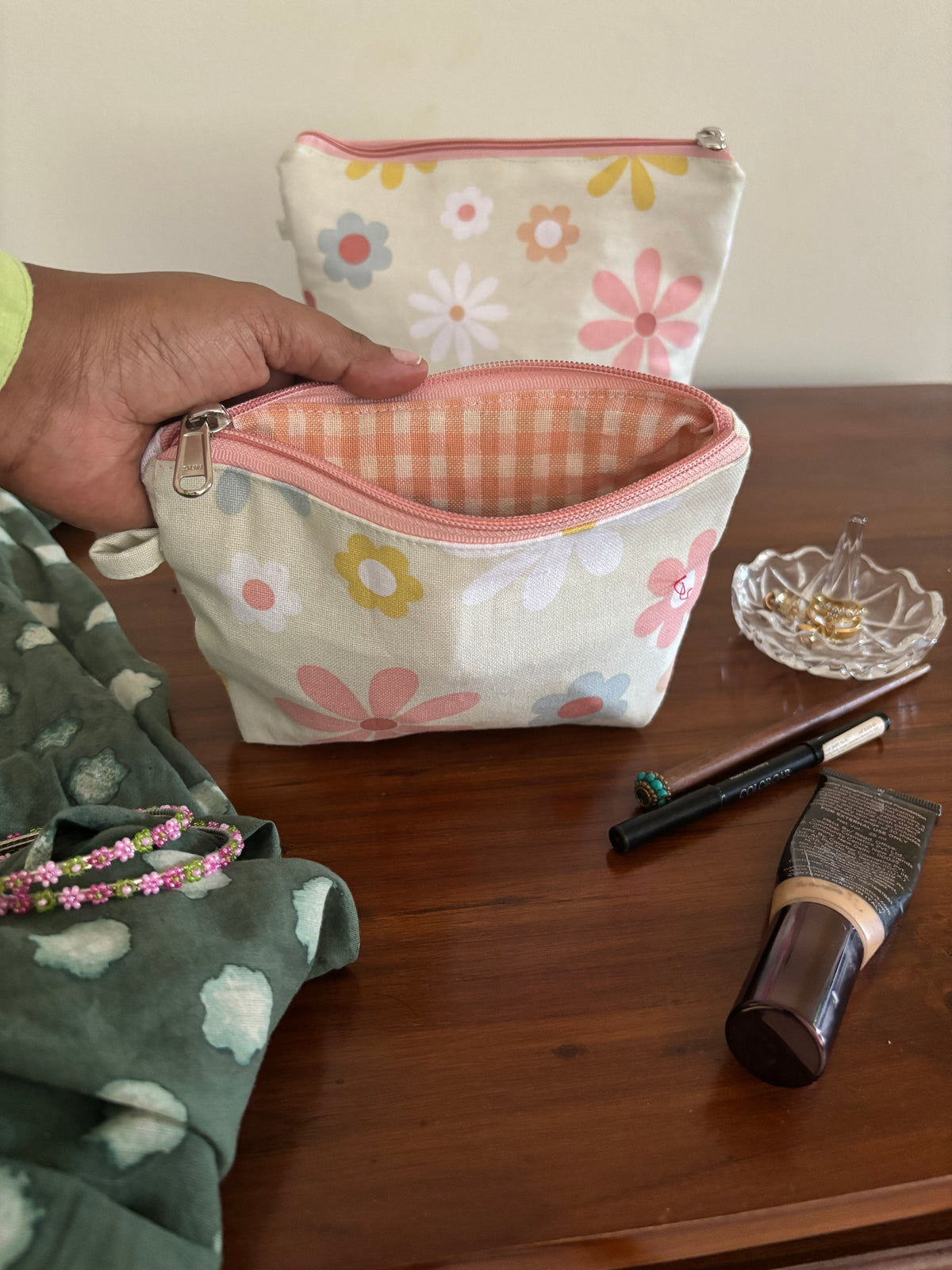 Hand holding a floral-patterned makeup bag with cosmetic items on a wooden surface.